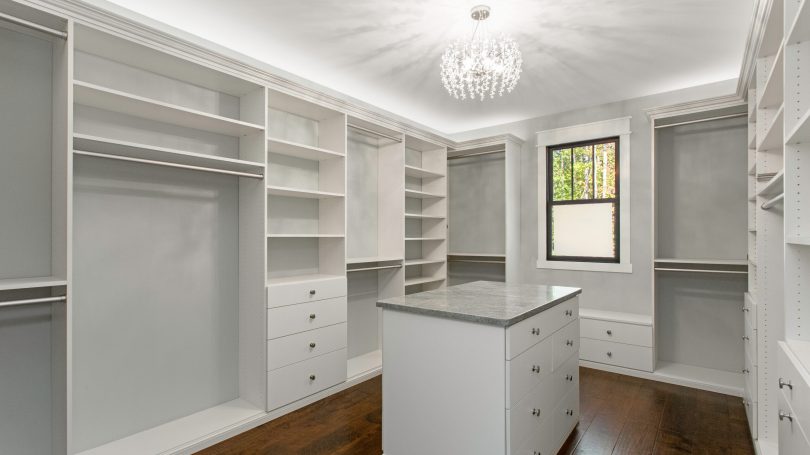 A spacious and luxurious walk-in closet featuring white shelving units and drawers, centered around a stylish island counter beneath a beautiful crystal chandelier.